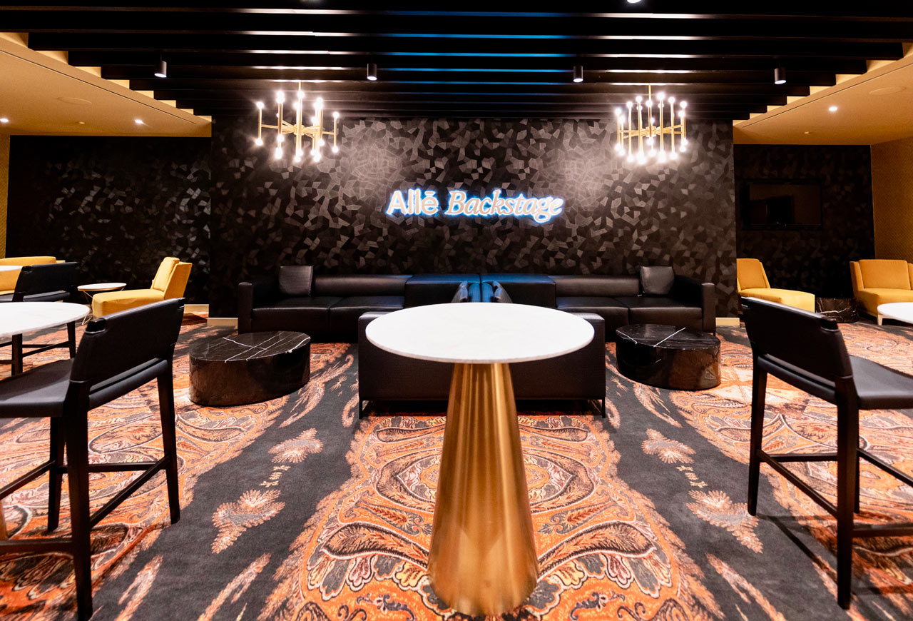 A modern lounge area with black and gold decor, featuring a neon sign that reads "Allē Backstage," black sofas, marble tables, and gold-accented furniture.